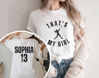 May include: A white t-shirt featuring a black graphic of a softball player swinging a bat with the text THAT'S MY GIRL. A circular inset shows the back of the same white t-shirt with the name SOPHIA and the number 13 printed in black.