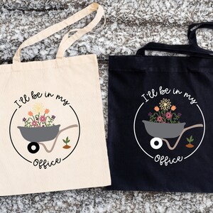 May include: Two tote bags with a white background and a black background. Both bags have a graphic of a wheelbarrow filled with flowers and the text "I'll be in my office".