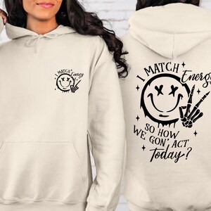 Puede incluir: Una sudadera con capucha de color crema con una cara sonriente negra y un gr&aacute;fico de mano de esqueleto en la parte delantera y el texto "I Match Energy So How We Gon' Act Today?" en la parte posterior.