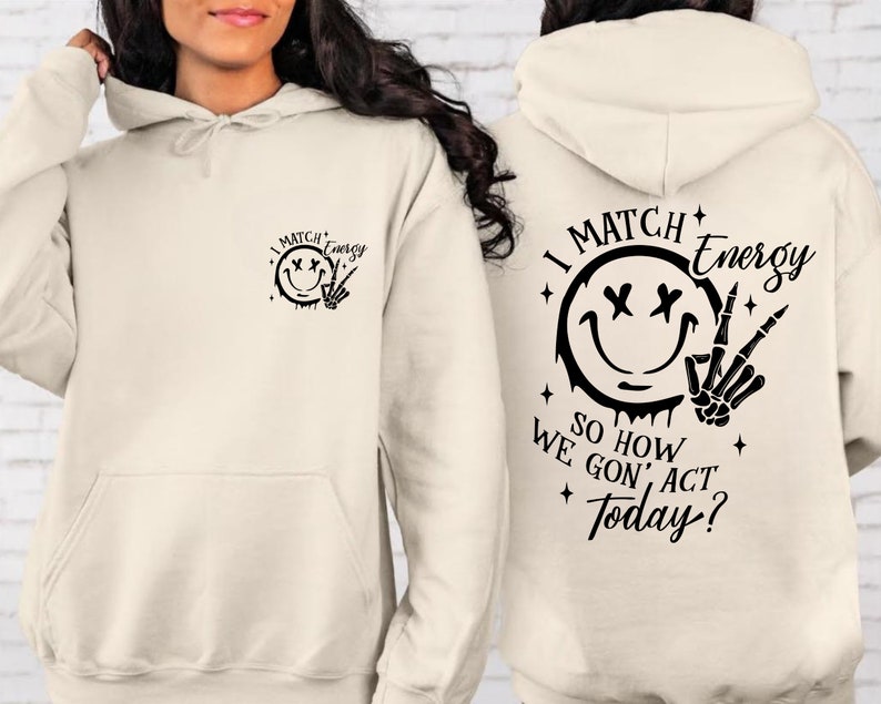 Puede incluir: Una sudadera con capucha de color crema con una cara sonriente negra y un gr&aacute;fico de mano de esqueleto en la parte delantera y el texto "I Match Energy So How We Gon' Act Today?" en la parte posterior.