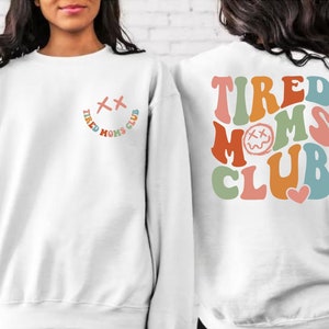 Puede incluir: Sudadera blanca con un gr&aacute;fico colorido de "Tired Moms Club" en la espalda y una versi&oacute;n m&aacute;s peque&ntilde;a del gr&aacute;fico en la parte delantera. El gr&aacute;fico presenta una cara sonriente con los ojos cruzados y un coraz&oacute;n.