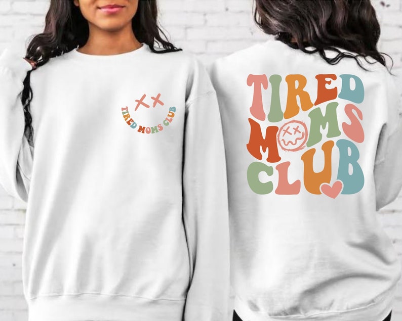 Puede incluir: Sudadera blanca con un gr&aacute;fico colorido de "Tired Moms Club" en la espalda y una versi&oacute;n m&aacute;s peque&ntilde;a del gr&aacute;fico en la parte delantera. El gr&aacute;fico presenta una cara sonriente con los ojos cruzados y un coraz&oacute;n.