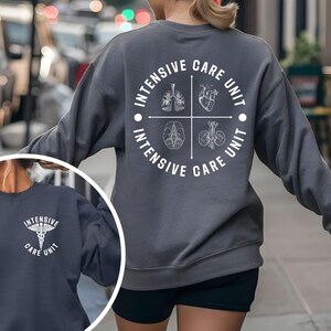 May include: A dark gray sweatshirt with the words "INTENSIVE CARE UNIT" in a circular design on the back. The design includes illustrations of lungs, a heart, a brain, and kidneys. A smaller image shows a similar sweatshirt with a medical symbol.