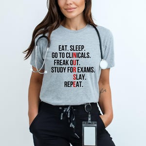 May include: A light gray t-shirt with the text "EAT. SLEEP. GO TO CLINICALS. FREAK OUT. STUDY FOR EXAMS. SLAY. REPEAT." in black and red lettering. A stethoscope hangs around the neck.
