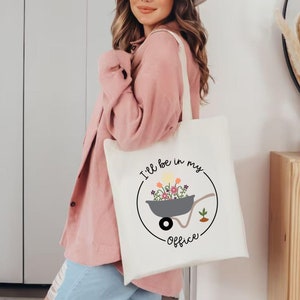 May include: White tote bag with a black circle design featuring a wheelbarrow filled with colorful flowers and the text "I'll be in my office".