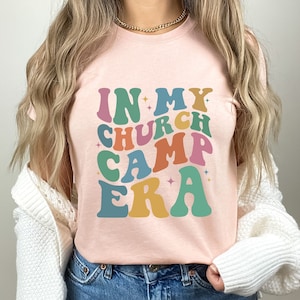 K&ouml;nnte beinhalten: Ein pinkfarbenes T-Shirt mit dem Text "IN MY CHURCH CAMP ERA" in bunten, Retro-Lettern.