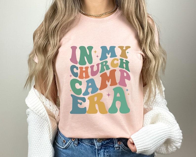 K&ouml;nnte beinhalten: Ein pinkfarbenes T-Shirt mit dem Text "IN MY CHURCH CAMP ERA" in bunten, Retro-Lettern.