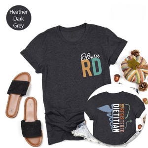 Puede incluir: Camiseta gris oscuro jaspeado con la inscripci&oacute;n "Olivia RD" en la parte delantera. La camiseta tiene cuello redondo y mangas cortas. Una imagen m&aacute;s peque&ntilde;a de la camiseta muestra la espalda con "Registered Dietitian" y un s&iacute;mbolo m&eacute;dico.