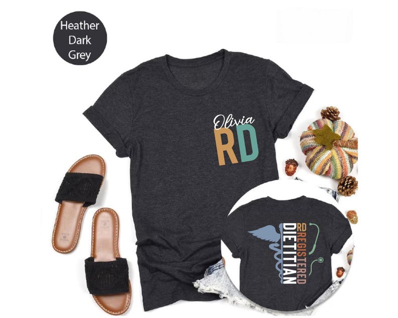 Puede incluir: Camiseta gris oscuro jaspeado con la inscripci&oacute;n "Olivia RD" en la parte delantera. La camiseta tiene cuello redondo y mangas cortas. Una imagen m&aacute;s peque&ntilde;a de la camiseta muestra la espalda con "Registered Dietitian" y un s&iacute;mbolo m&eacute;dico.
