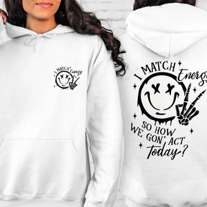 Puede incluir: Sudadera con capucha blanca con una cara sonriente negra y un gr&aacute;fico de mano de esqueleto. El texto "I Match Energy So How We Gon' Act Today?" est&aacute; impreso en la sudadera.