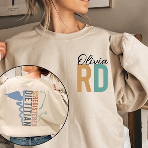 May include: Beige sweatshirt with the name "Olivia" and "RD" in black, tan, and teal. The back features a Registered Dietitian design with a caduceus, stethoscope, and text. A comfortable and stylish garment.
