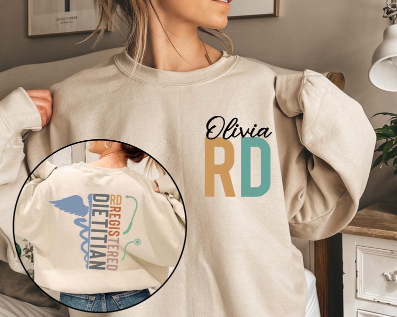 May include: Beige sweatshirt with the name "Olivia" and "RD" in black, tan, and teal. The back features a Registered Dietitian design with a caduceus, stethoscope, and text. A comfortable and stylish garment.