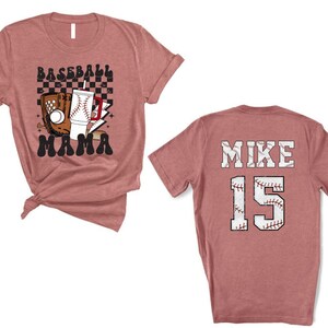 May include: A dusty rose baseball t-shirt with a graphic on the front that says "Baseball Mama". The graphic includes a baseball glove, a baseball, and a drink. The back of the shirt says "MIKE 15".