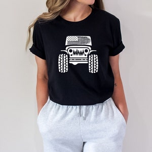 May include: Black t-shirt with a white graphic of a Jeep with an American flag on the hood and trees in the grill. The Jeep has large tires.