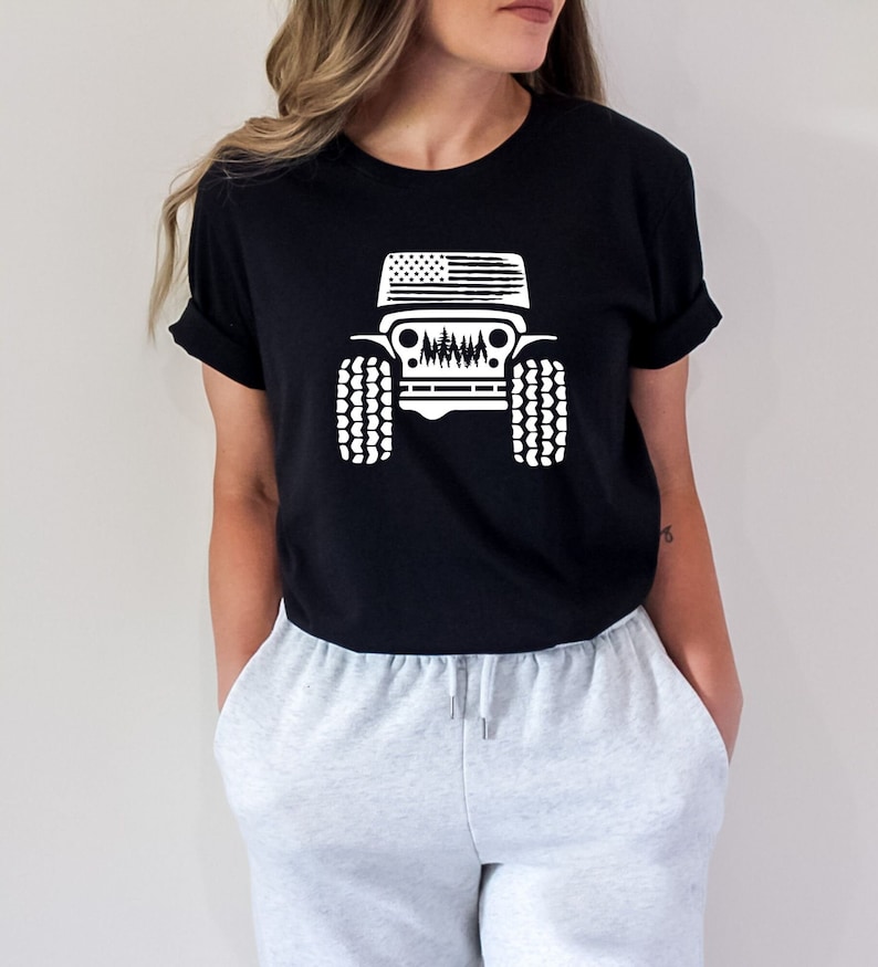 May include: Black t-shirt with a white graphic of a Jeep with an American flag on the hood and trees in the grill. The Jeep has large tires.