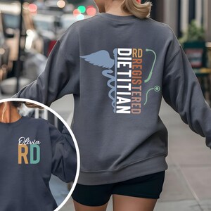 May include: A gray sweatshirt featuring the text "RD REGISTERED DIETITIAN" with a caduceus symbol and a stethoscope graphic. The sweatshirt also has a smaller image with the name "Olivia" and the letters "RD".