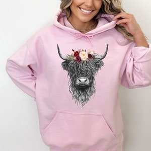 Puede incluir: Una sudadera con capucha rosa con una ilustraci&oacute;n en blanco y negro de una vaca de las tierras altas con una corona de flores. Las flores son rojas, rosas y blancas.