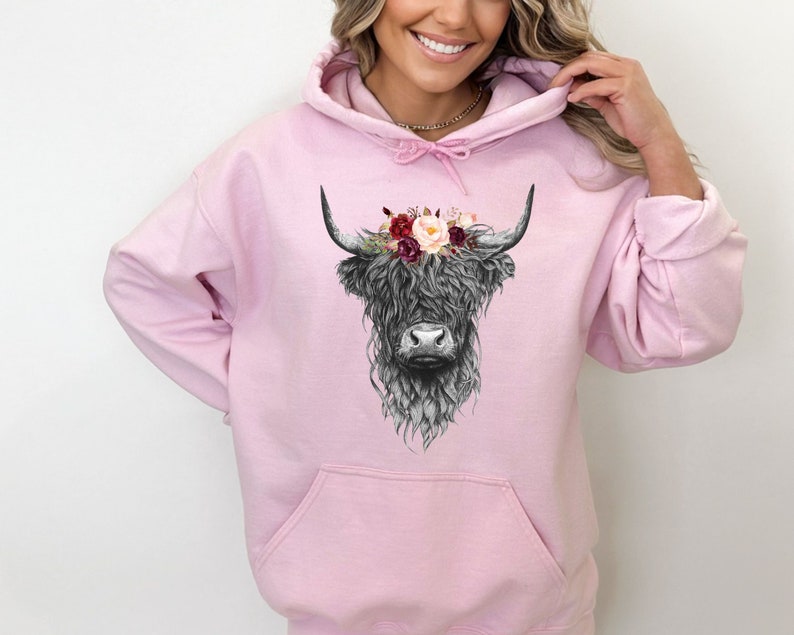 Puede incluir: Una sudadera con capucha rosa con una ilustraci&oacute;n en blanco y negro de una vaca de las tierras altas con una corona de flores. Las flores son rojas, rosas y blancas.