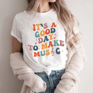 May include: A white short-sleeved t-shirt featuring the multi-colored text IT'S A GOOD DAY TO MAKE MUSIC, with the letter I in MUSIC replaced by a treble clef symbol. The graphic includes small star and musical note accents in red, blue, and orange.