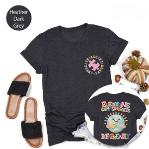 May include: Two heather dark grey t-shirts with a graphic design. The first shirt has a pink puzzle piece inside a circle with the words "Advocate, Accept, Adapt". The second shirt has a cartoon earth holding a puzzle piece with the words "Everyone Communicates Differently".