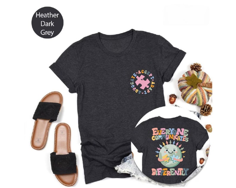 May include: Two heather dark grey t-shirts with a graphic design. The first shirt has a pink puzzle piece inside a circle with the words "Advocate, Accept, Adapt". The second shirt has a cartoon earth holding a puzzle piece with the words "Everyone Communicates Differently".