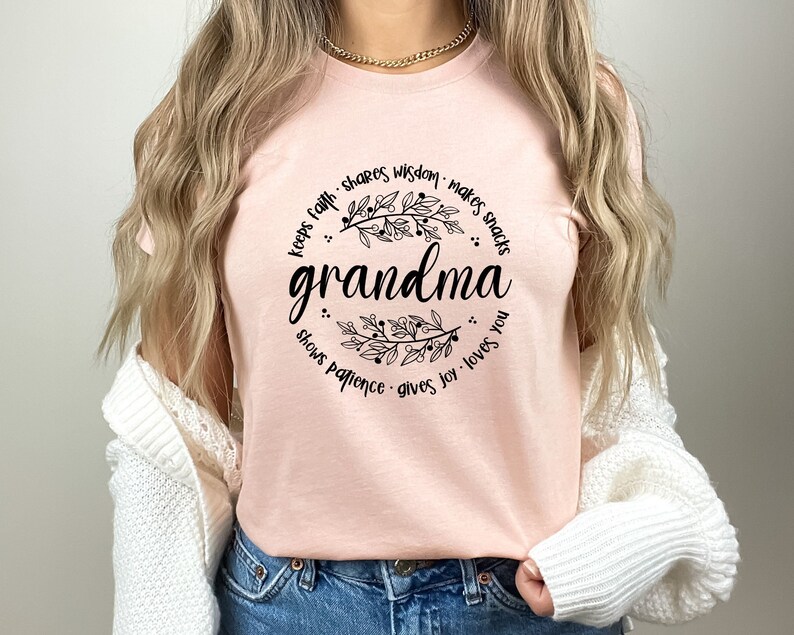 Op de afbeelding: Een lichtroze t-shirt met het woord "grandma" in zwart script, omcirkeld door een bloemenkrans. De tekst rond de krans luidt "keeps faith, shares wisdom, makes snacks, shows patience, gives joy, loves you."