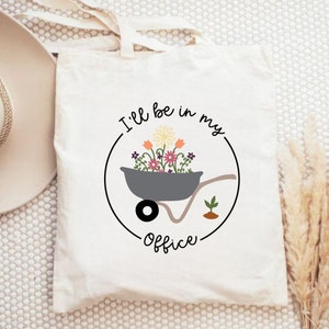 May include: White canvas tote bag with a black circle design. The circle contains a gray wheelbarrow filled with colorful flowers and the text "I'll be in my office".