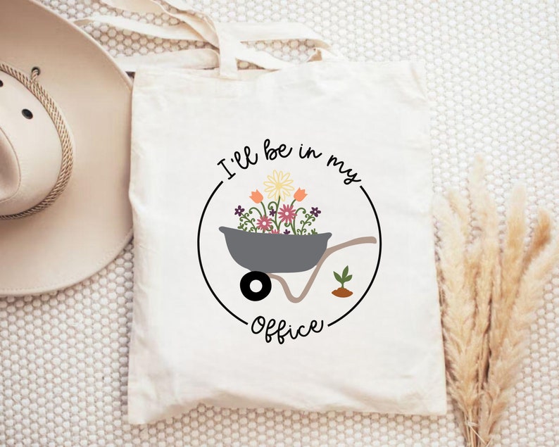 May include: White canvas tote bag with a black circle design. The circle contains a gray wheelbarrow filled with colorful flowers and the text "I'll be in my office".
