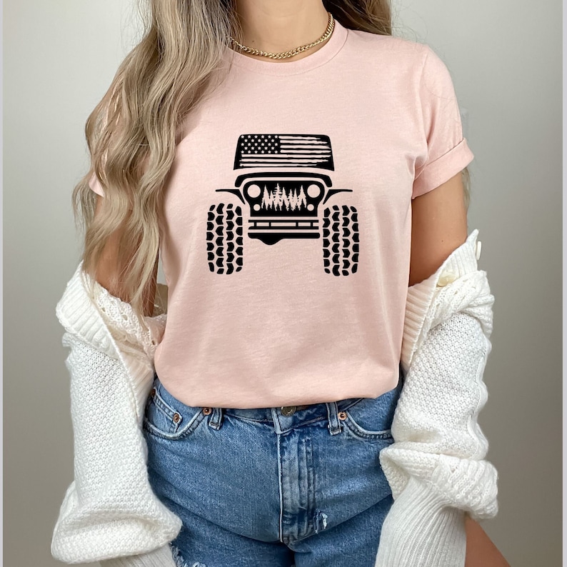 May include: A pink t-shirt with a black graphic of a Jeep with an American flag on the hood and trees in the grill. The Jeep has black tires.