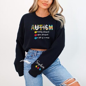 May include: Black cropped sweatshirt with the text "AUTISM mama totally blessed often stressed a bit of a mess" in colorful puzzle piece designs. The sweatshirt has a personalized name on the sleeve.