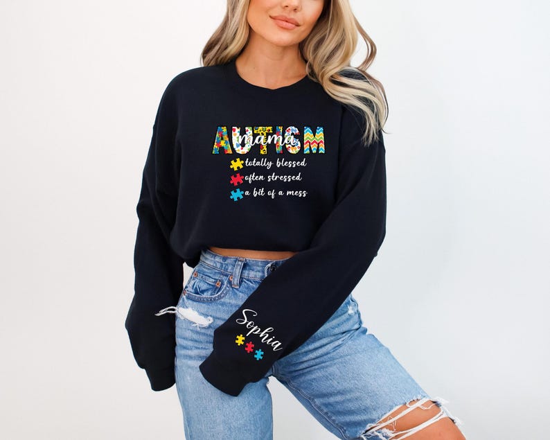 May include: Black cropped sweatshirt with the text "AUTISM mama totally blessed often stressed a bit of a mess" in colorful puzzle piece designs. The sweatshirt has a personalized name on the sleeve.