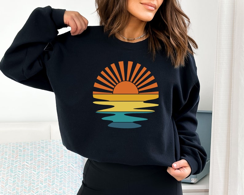 May include: Black sweatshirt featuring a retro sunset design. The graphic includes a sun with orange and yellow rays, reflected in layered bands of yellow, teal, and blue. A casual, comfortable top for everyday wear.