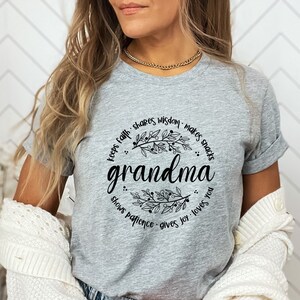 Op de afbeelding: Een heather grijze t-shirt met het woord "grandma" in een sierlijk lettertype, omcirkeld door een bloemenkrans. De krans bevat zinnen als "keeps faith", "shares wisdom" en "gives joy". De mouwen van de shirt zijn opgerold.