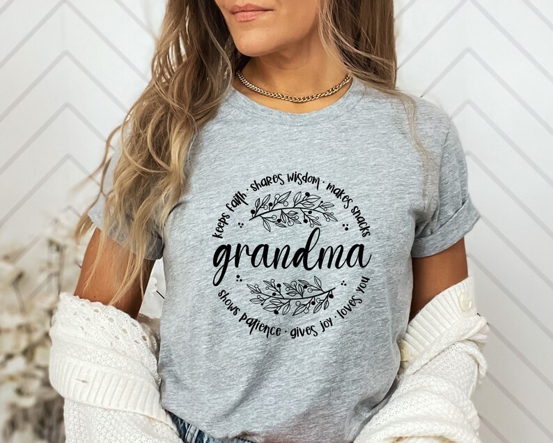 Op de afbeelding: Een heather grijze t-shirt met het woord "grandma" in een sierlijk lettertype, omcirkeld door een bloemenkrans. De krans bevat zinnen als "keeps faith", "shares wisdom" en "gives joy". De mouwen van de shirt zijn opgerold.