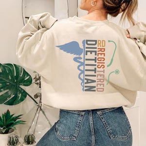 May include: Cream-colored sweatshirt with the words "RD REGISTERED DIETITIAN" and a caduceus symbol in blue, teal, and maroon. A stethoscope graphic is also present. The sweatshirt is a casual garment.
