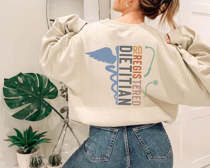 May include: Cream-colored sweatshirt with the words "RD REGISTERED DIETITIAN" and a caduceus symbol in blue, teal, and maroon. A stethoscope graphic is also present. The sweatshirt is a casual garment.
