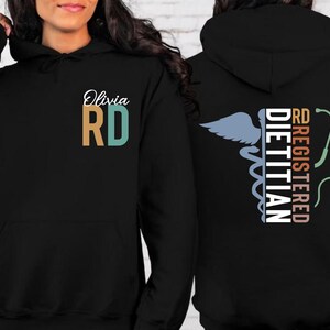 May include: Black hoodie with the text "Olivia RD" on the front, with the letters in teal and tan. The back of the hoodie features a caduceus symbol, a stethoscope, and the words "RD REGISTERED DIETITIAN".