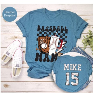 May include: A teal t-shirt with a baseball-themed graphic that says "Baseball Mama." The design includes a baseball glove, a baseball, and a drink with a baseball design. A second shirt shows "Mike 15" on the back. White sneakers are visible.