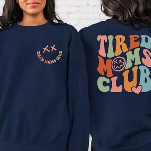 Puede incluir: Sudadera azul marino con un gr&aacute;fico colorido de "Tired Moms Club" en la espalda y una versi&oacute;n m&aacute;s peque&ntilde;a del gr&aacute;fico en la parte delantera. El gr&aacute;fico presenta una cara sonriente con X para los ojos.