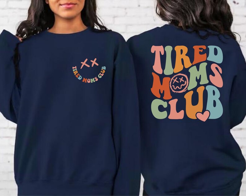 Puede incluir: Sudadera azul marino con un gr&aacute;fico colorido de "Tired Moms Club" en la espalda y una versi&oacute;n m&aacute;s peque&ntilde;a del gr&aacute;fico en la parte delantera. El gr&aacute;fico presenta una cara sonriente con X para los ojos.