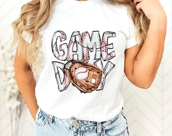 May include: A white short-sleeved t-shirt featuring the text GAME DAY in a stylized font with a red baseball stitch pattern. The letter A in DAY is replaced by a brown leather baseball glove holding a white baseball with red stitching.