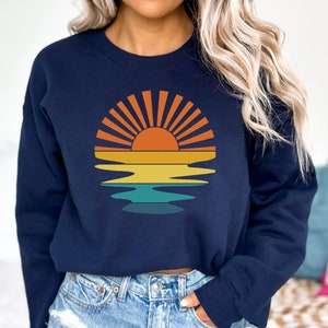 May include: Navy blue sweatshirt featuring a retro sunset design. The graphic includes a sun with orange rays, a yellow and orange gradient, and a teal water reflection. A casual, comfortable top for everyday wear.