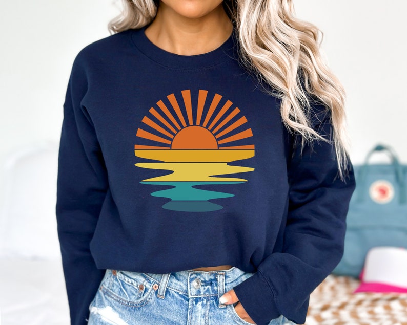 May include: Navy blue sweatshirt featuring a retro sunset design. The graphic includes a sun with orange rays, a yellow and orange gradient, and a teal water reflection. A casual, comfortable top for everyday wear.