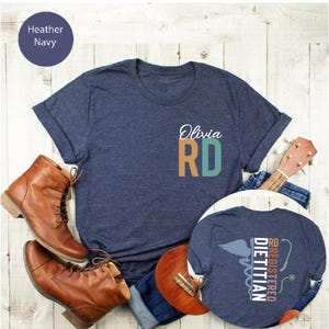 Puede incluir: Camiseta azul marino con el nombre "Olivia" y "RD" en verde azulado y naranja. La camiseta se muestra con botas marrones, una guitarra y vaqueros. Una segunda camiseta muestra el texto "RD Registered Dietitian" con un s&iacute;mbolo m&eacute;dico.