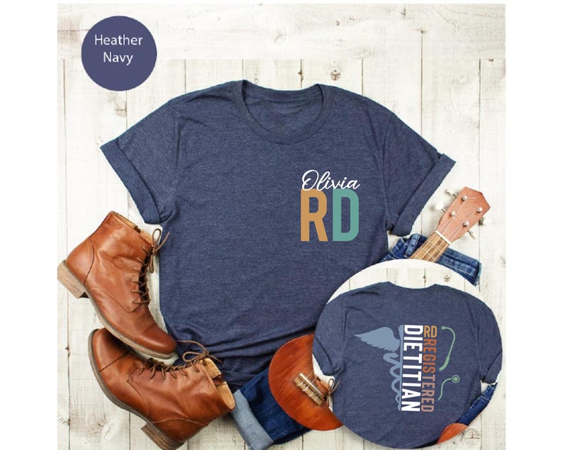 Puede incluir: Camiseta azul marino con el nombre "Olivia" y "RD" en verde azulado y naranja. La camiseta se muestra con botas marrones, una guitarra y vaqueros. Una segunda camiseta muestra el texto "RD Registered Dietitian" con un s&iacute;mbolo m&eacute;dico.