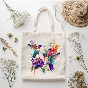 May include: A white tote bag with a colorful watercolor design of three hummingbirds and flowers. The hummingbirds are flying and have vibrant blue, green, and purple feathers. The flowers are pink, purple, and orange.