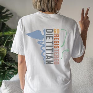 Puede incluir: Camiseta blanca con un s&iacute;mbolo m&eacute;dico y las palabras "RD REGISTERED DIETITIAN" en varios colores. La camiseta presenta un s&iacute;mbolo de caduceo azul, un estetoscopio y texto en naranja y negro.