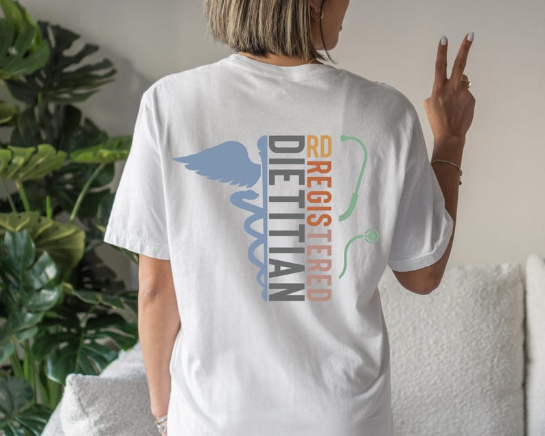 Puede incluir: Camiseta blanca con un s&iacute;mbolo m&eacute;dico y las palabras "RD REGISTERED DIETITIAN" en varios colores. La camiseta presenta un s&iacute;mbolo de caduceo azul, un estetoscopio y texto en naranja y negro.