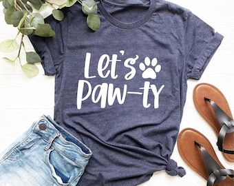 Puede incluir: Una camiseta de manga corta en azul marino jaspeado con el texto blanco Let's Paw-ty y un gráfico de huella. La prenda se muestra junto a unos pantalones cortos de mezclilla azul claro y sandalias marrones, con hojas verdes arriba.
