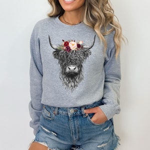 Puede incluir: Una sudadera gris con una ilustraci&oacute;n en blanco y negro de una vaca de las tierras altas con una corona de flores. Las flores son rojas, rosas y blancas.
